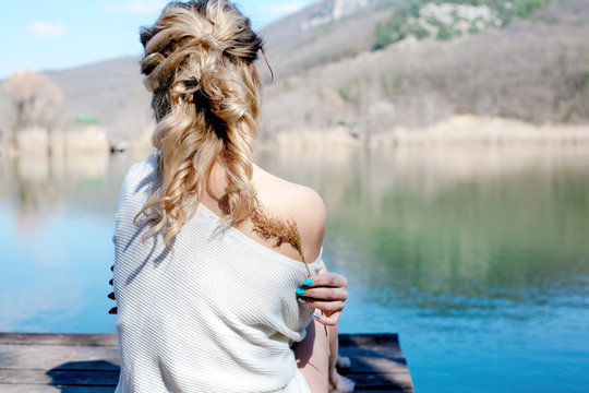 Rear view of the back of woman relax in the wood piar near the lake. Female hands hug herself and look to wildlife of the nature. model wearin bohemian accessories and oversize sweater.