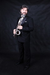 Full length profile shot of male artist musician in a classic black suit, tailcoat, statuesque in a bow tie with a beard plays music on a gold saxophone.black background