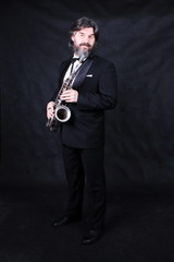 Naklejka premium Full length profile shot of male artist musician in a classic black suit, tailcoat, statuesque in a bow tie with a beard plays music on a gold saxophone.black background