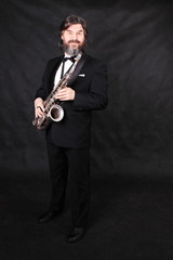 Full length profile shot of male artist musician in a classic black suit, tailcoat, statuesque in a bow tie with a beard plays music on a gold saxophone.black background