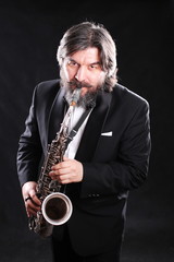 Fototapeta na wymiar A male artist musician in a classic black suit, tailcoat, statuesque in a bow tie with a beard plays music on a gold saxophone.black background
