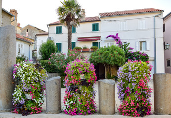 Fototapeta na wymiar Krk, Croatia - Three large bulbs with colorful flowers between four dilapidated stone columns, a palm tree, trees with flowers and a white building with a brown roof in the background in the summer.