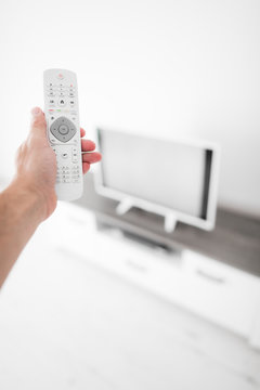 Hand holding TV remote controller with a modern television set.