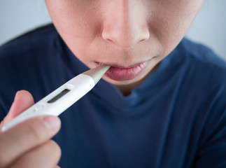 Young Asian woman has a cold and high fever, measuring temperature her body by using digital thermometer in the living room. Health care and medical concept.