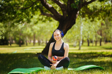 Beautiful girl in sportswear sitting on a green mat in the park with a glass of water. Doing yoga with headphones. Listens to music during sports