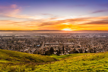 Sunset view of residential and industrial areas in East San Francisco Bay Area; green hills visible in the foreground; Hayward, California
