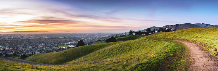 Fototapeta na wymiar Sunset view of hiking trail on the verdant hills of East San Francisco Bay Area; the city of Hayward and the bay visible in the valley; California