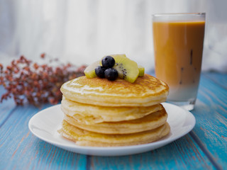 Stack of pancakes with topping, kiwi and blueberry.placed in a white plate on a blue wooden table.Eat with milk tea in the glass.