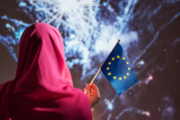 Muslim woman in a scarf holding flag of EU (Europe) during fireworks at night.
