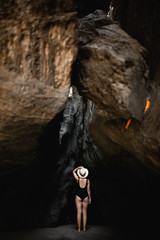 young girl in a black adjacent swimsuit and a straw hat stands on the sand in a narrow cave and looks into the gorge - 316954292