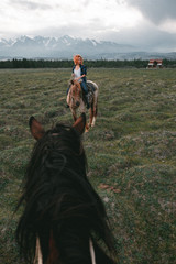 on a green field, a red-haired girl sits astride a horse, holds her by the reins and looks to the side, against a background of snowy mountain ranges - 316956022