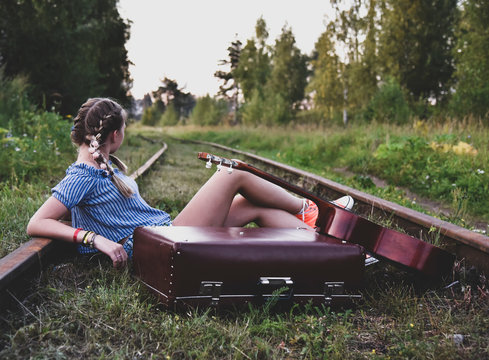 A teenage girl is sitting on the rails with a guitar and an old suitcase