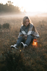 vertical shot of a young girl with blond hair in a warm woolen sweater wrapped in a plaid, she sits in a field with tall dry grass at dawn with a kerasin lamp - 316959205