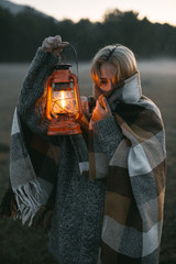 vertical shot of a young girl with blond hair in a warm woolen sweater wrapped in a plaid, she stands at dawn with a kerasin lamp in her hands - 316959664