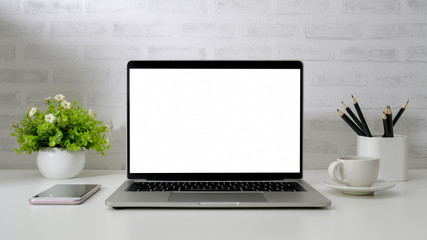 Close up view of workspace with blank screen laptop, smartphone, pencils, coffee cup and tree pot on white desk with  brick wall