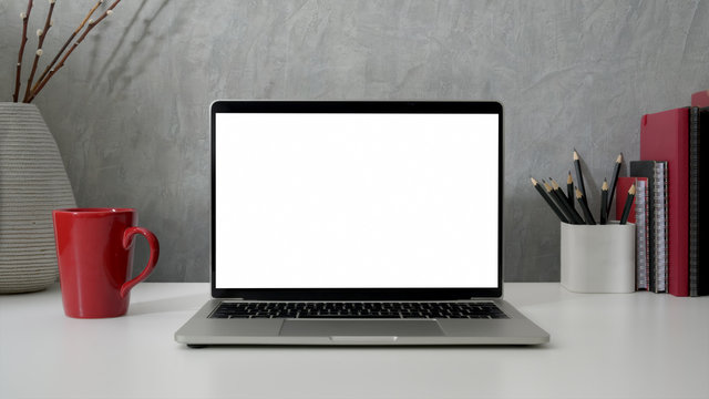 Close up view of workspace with blank screen laptop, office supplies and red coffee cup on white table with grey loft wall