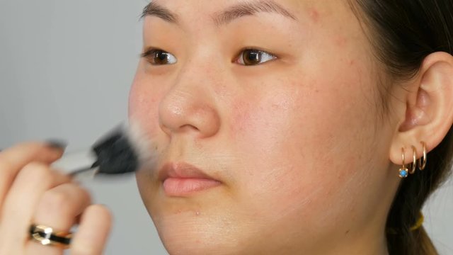 Professional makeup artist applies foundation concealer or highlighter to Asian Korean model's face with a special brush