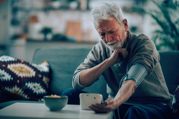 Old man measuring blood pressure. Sad senior man in living room. 