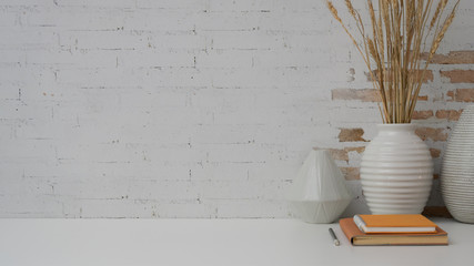 Close up view of workplace with copy space, notebooks and ceramic vase on white desk with brick wall