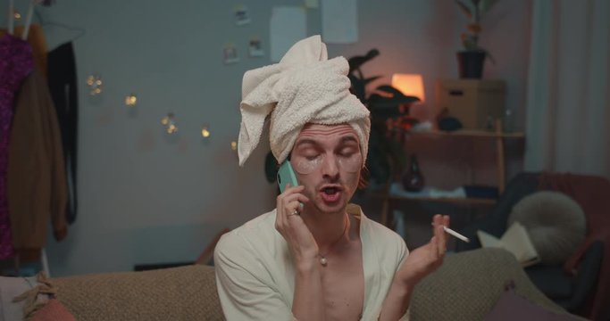 Close up of young angry transgender smoking cigarette and communicating on smartphone. Man transin home wear looking disaffected sitting on sofa and yelling with somebody on phone.