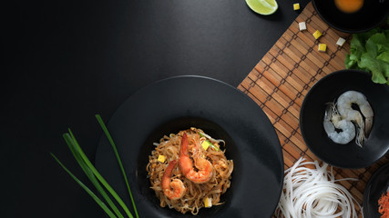Overhead shot of Pad Thai, stir fly of Thai noodle with shrimp and egg in black ceramic plate on bamboo placemat with ingredients