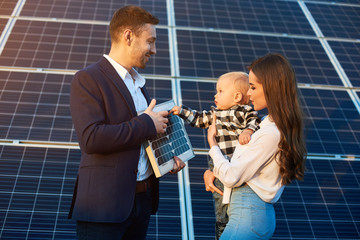 Cheerful kid in his mother arms examines the solar battery that dad holds, against background solar...