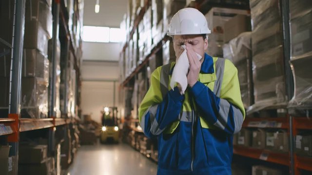 Portrait of young sick warehouse worker blowing his nose while standing in industrial storage