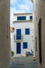 Cadaqués, Catalonia / Spain - November 30th, 2019: Narrow street in the old village with a white facade house with blue shutters and a blue scooter