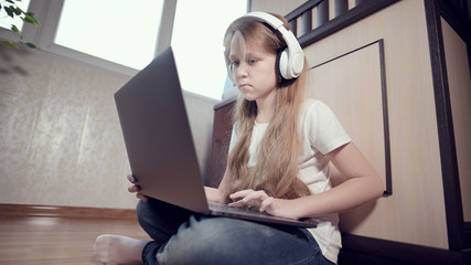 A smart little girl of seven years old in white headphones with a laptop in her hands is pushing on the floor in her room. The young generation on the Internet and IT technology