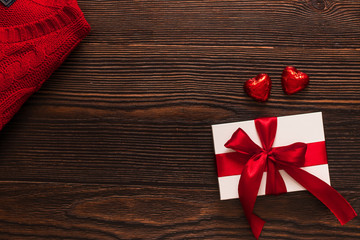 White present with a red ribbon, red sweater and two chocolate red hearts isolated on a dark wooden background. Top side view of flatlay. Valentines day and Christmas concept. Copyspace.