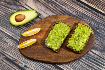 Avocado paste is spread on rye bread with seeds. Near slices of lemon and half an avocado. Appetizing avocado and guacamole toast. Healthy breakfast concept. Wooden background, flat lay.