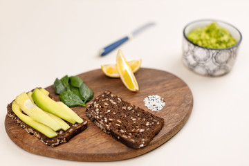 Close up photo of ingredients for avocado toast. Dark bread with seeds, basil leaves, lemon slices and sesame seeds on wooden board. Guacamole in bowl. Mexican cuisine concept. Vegetarian sandwiches.