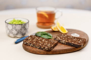 Whole wheat bread, basil leaves, lemon slices and sesame seeds on wooden board. Avocado paste and guacamole in bowl. Ingredients for avocado toast. Selective focus, blurred background. Mexican cuisine