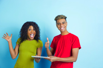 Holding tablet, thumbs up. Young emotional african-american man and woman in colorful casual clothes on blue background. Beautiful couple. Concept of human emotions, facial expession, relations, ad.