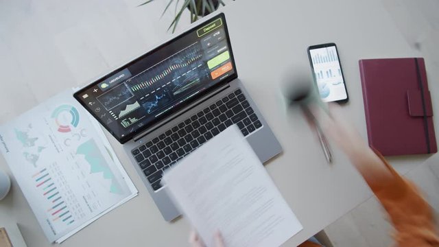 Timelapse top shot of female investment analyst sitting at desk in office, writing in notebook, using smartphone, analyzing diagrams and graphs and typing on laptop with currency trading software