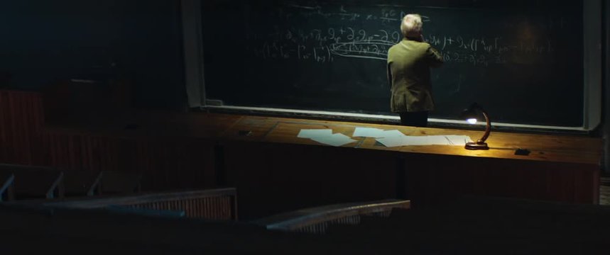 Aged professor or scientist is working on solving a complex equation or formula late at night. University auditorium, table lamp shines. Anamorphic lens