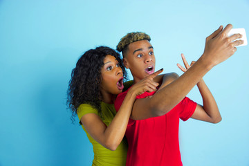 Taking selfie or vlog together. Young emotional african-american man and woman in colorful clothes on blue background. Beautiful couple. Concept of human emotions, facial expession, relations, ad.