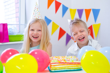 Fototapeta na wymiar Two blonde caucasian kids boy and girl having fun and laughing at birthday party. Colorful background with balloons and birthday rainbow cake