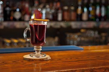 Red cocktail on bar counter. Mulled wine with fruits and cinnamon on the background of bottles with alcohol. Glass with booze on a wooden board. Cinnamon burns and smokes.