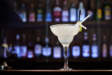 White cocktail on bar counter. Drink with martini, gin, tequila and lime on a background of bottles with alcohol. Glass with booze on a wooden board. Blue background with ultraviolet illumination.
