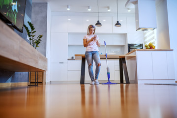 Beautiful Caucasian blond housewife dressed casual leaning on dining table and using smart phone for sending message. Next to her is mop.