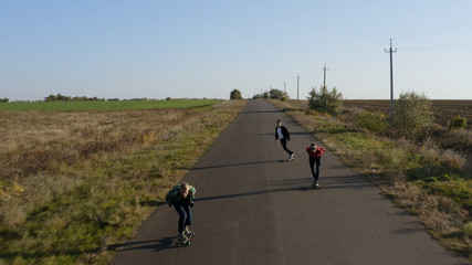 Aerial view of three hipster men skateboarding on road in meadow at sunset drone