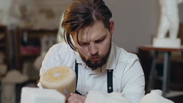 Slow motion of good-looking focused on his work bearded young sculptor which giving the desired shape to the intended sculpture with hammer and chisel