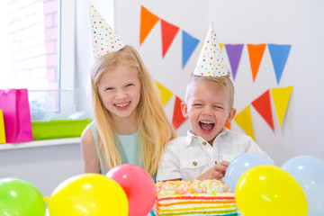 Fototapeta na wymiar Two blonde caucasian kids boy and girl having fun and laughing at birthday party. Colorful background with balloons and birthday rainbow cake