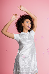 attractive happy african american girl with dental braces, with silver glitter eyeshadows and purple lips dancing in paillettes dress, isolated on pink with confetti