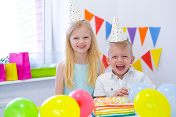Fototapeta na wymiar Two blonde caucasian kids boy and girl in birthday hats looking at camera and smiling at birthday party. Colorful background with balloons and birthday rainbow cake