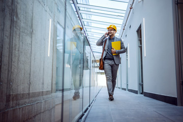 Full length of smiling caucasian hardworking businessman with folder in hands going to a meeting while talking on the phone. Future shopping mall interior.