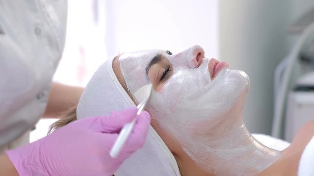 Cosmetologist is applying white mask on woman client face in beauty clinic. Portrait of woman, side view. Beautician making beauty facial skincare procedure to patient. Beauty industry concept.