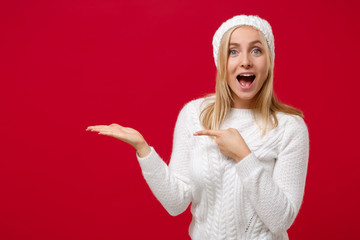 Amazed young woman in white sweater hat isolated on red background in studio. Healthy fashion lifestyle cold season concept. Mock up copy space. Pointing hand, index finger aside, keeping mouth open.