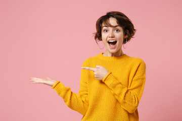 Cheerful excited young brunette woman girl in yellow sweater posing isolated on pastel pink background studio portrait. People lifestyle concept. Mock up copy space. Pointing index finger, hand aside.